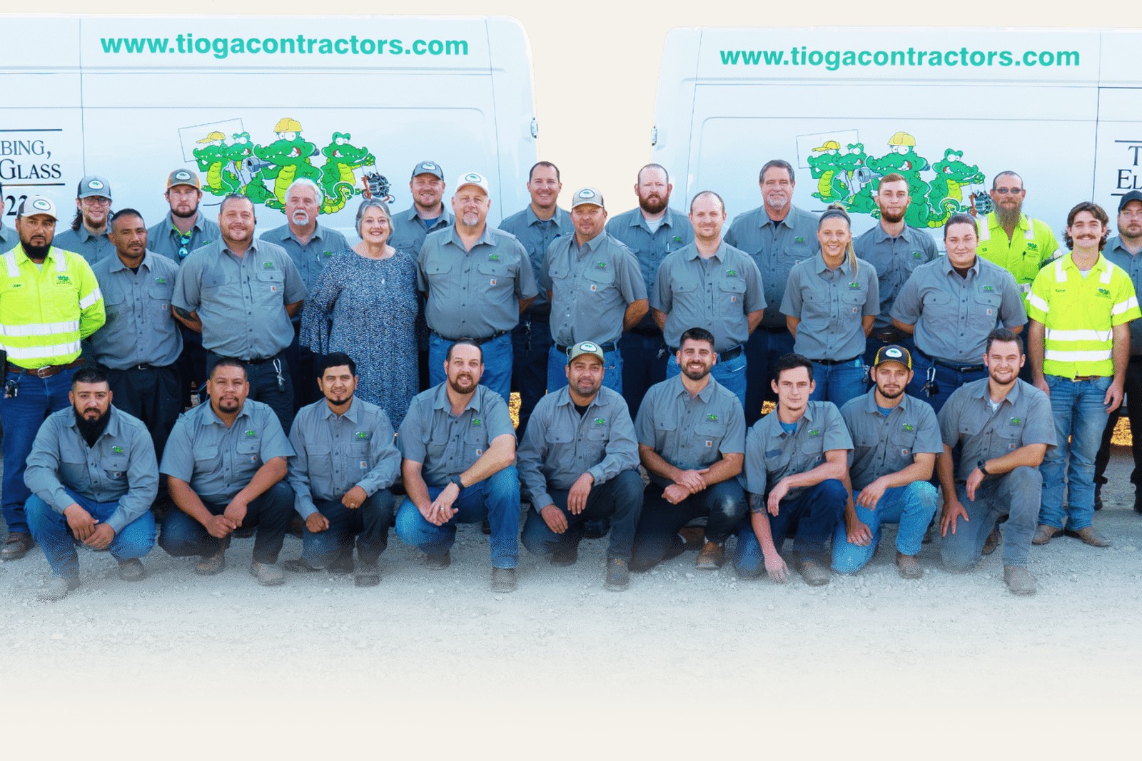 The full Tioga Plumbing crew in gray work uniforms lined up in front of two branded service vans at the Hurst, TX shop