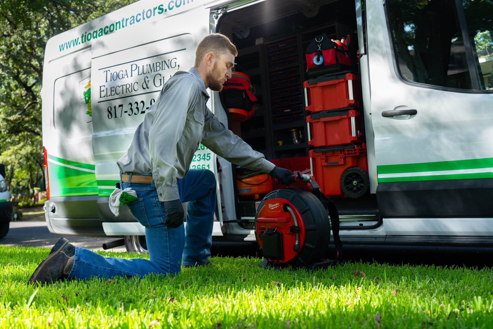 Tioga Plumbing technician kneeling on a DFW front lawn, loading a drain-cleaning machine into a branded service van