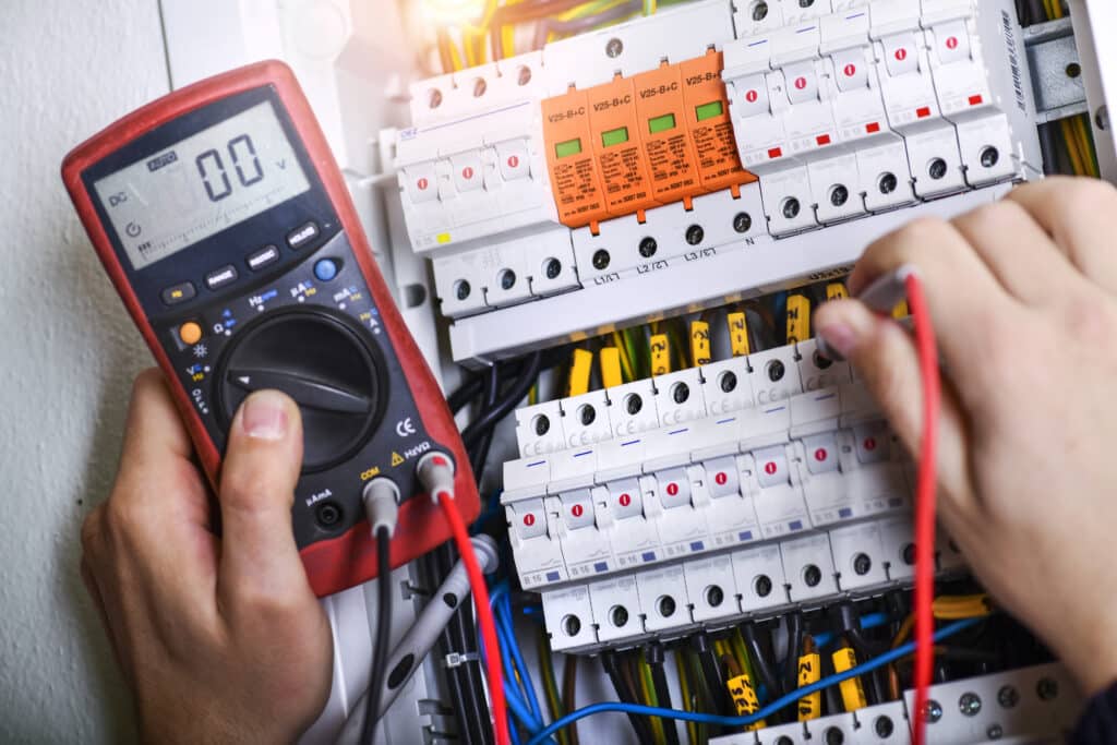 The electrician is installing electrical cable wires, a fuse switch box, and surge protection devices. A multimeter is in the electrician's hands.